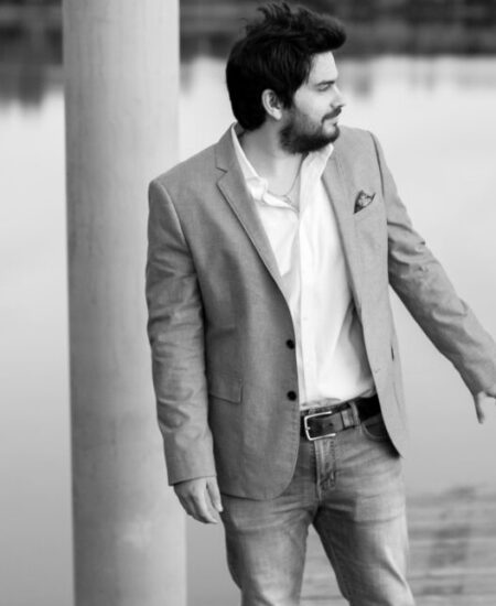 A man with dark hair and a beard stands on a wooden dock wearing a light-colored blazer and jeans. He looks to his right with a neutral expression. A large, vertical pillar and a body of water are in the background. The image is in black and white.